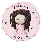Circular logo for EmmaS Dolls featuring a handmade crochet doll with curly brown hair and a pink dress. The doll is centered on a soft pink background, framed by a delicate woody wreath with small green leaves and tiny mint-colored hearts. The text 'EMMAS DOLLS' is written in a clean, black serif font arc above and below the doll.